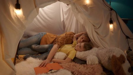 Téléchargez les photos : Papa et fils dormant blottis sur un grand jouet en peluche, dans une tente dans le salon. L'homme s'est endormi sans lâcher le livre. Portrait de l'homme endormi et garçon gros plan - en image libre de droit