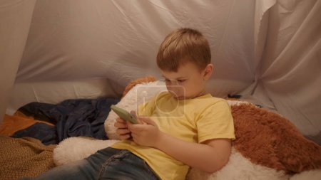 Téléchargez les photos : Le garçon est allongé sur un gros jouet doux avec un smartphone dans les mains de près. Un petit garçon passe la soirée à jouer à un jeu en ligne sur un smartphone, couché dans une tente, qui est installé dans le salon - en image libre de droit