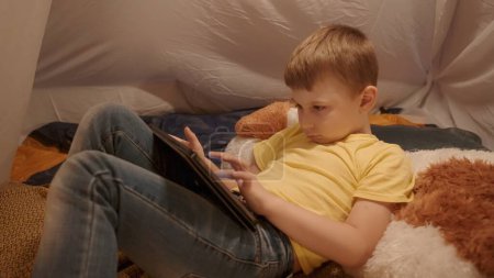 Foto de El niño está acostado en un gran juguete blando con una tableta de cerca. Un niño pasa la noche jugando un juego en línea en una tableta, acostado en una tienda de campaña, que se instala en la sala de estar - Imagen libre de derechos