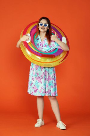 Photo for Travelling and vacation advertisement concept. Studio shot of female model. Woman in glasses with colorful inflatable pool tube over neck showing thumbs up, smiling, isolated on orange background. - Royalty Free Image