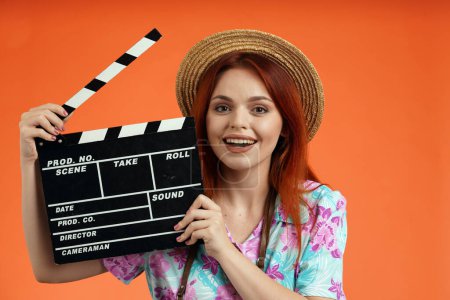Photo for Travelling advertisement concept. Studio shot of female model. Close up of happy woman in straw hat smiling and holding opened clapperboard looking at the camera, isolated on orange background. - Royalty Free Image