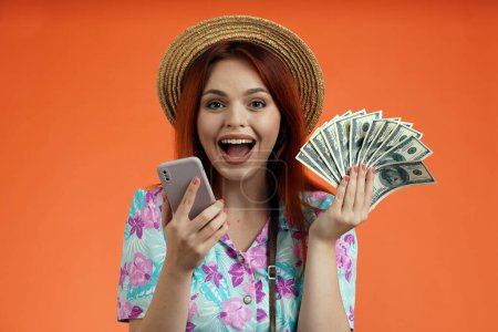 Photo for Travelling advertisement concept. Studio shot of female model. Close up of surprised woman in straw hat smiling, holding smartphone and fan of money bills at the camera, isolated on orange background. - Royalty Free Image