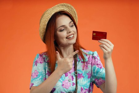 Photo for Travelling advertisement concept. Studio shot of female model. Close up of happy woman in straw hat smiling, holding bank credit card in hand and pointing at it, isolated on orange background. - Royalty Free Image