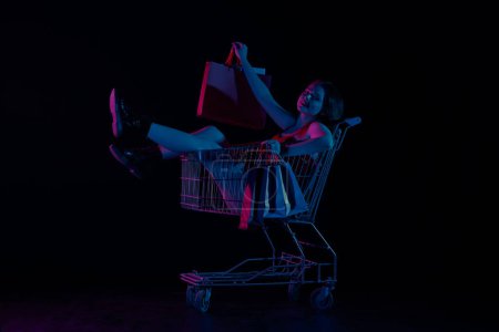 Photo for Black friday and seasons sale advertisement concept. Attractive woman sitting in shopping cart with hands full of shopping paper bags, looking at camera. Isolated on black background in neon light. - Royalty Free Image