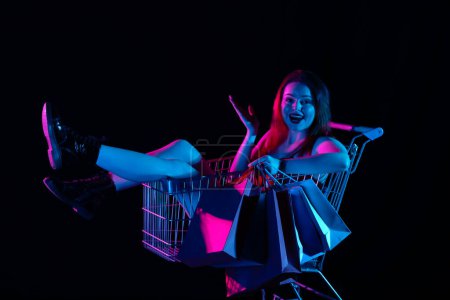 Photo for Black friday and seasons sale advertisement concept. Attractive woman sitting in shopping cart holding shopping bags, smiling amazed face expression Isolated on black background in neon light. - Royalty Free Image