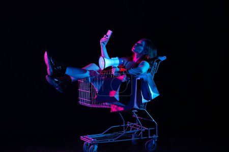 Photo for Black friday and seasons sale advertisement concept. Attractive woman sitting in shopping cart holding smartphone and megaphone, smiling taking selfie. Isolated on black background in neon light. - Royalty Free Image