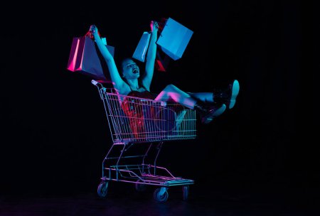 Photo for Black friday and seasons sale advertisement concept. Attractive woman sitting in shopping cart with hands up holding shopping bags, overjoyed face expression. Isolated on black background neon light. - Royalty Free Image