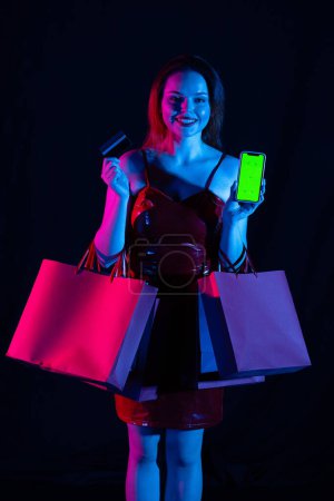 Photo for Black friday and seasons sale advertisement concept. Attractive woman holds credit card and smartphone with chroma key green screen, smiling. Advertising area. Isolated on black background neon light. - Royalty Free Image