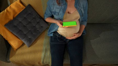 Photo for A close up shot shows a pregnant woman sitting on a sofa. She is holding a phone with a green screen in her hands. She examines him. Here can be your advertising. Top view. Medium shot. - Royalty Free Image