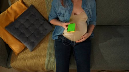 Téléchargez les photos : Un gros plan montre une femme enceinte assise sur un canapé. Elle tient un téléphone avec un écran vert dans ses mains. Elle l'examine. Voici peut être votre publicité. Vue de dessus. Plan moyen. - en image libre de droit