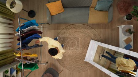 Photo for Shopping, leisure and lifestyle advertisement concept. Top view of living room. Man standing next to hanger with clothes ant looking through many colorful clothes on it, picking outfit. - Royalty Free Image