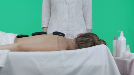 Photo for Body wellbeing creative concept. Beauty salon room, female on massage table with spa stones on the back, masseur standing near. Chroma key green screen background, advertising area workspace mockup. - Royalty Free Image