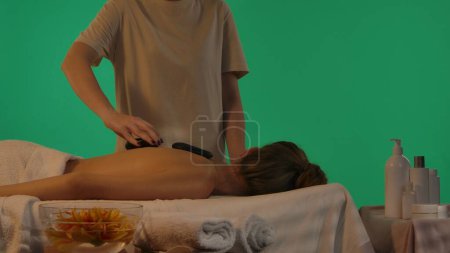 Photo for Body wellbeing creative concept. Spa salon, female on massage table, woman putting on black stones on the female model back. Chroma key green screen background, advertising area workspace mockup. - Royalty Free Image