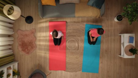 Foto de En la foto de arriba, una mujer y un niño están de pie en una habitación en una alfombra de fitness. Están demostrando una pose de yoga. La mujer le está enseñando a la chica cómo hacerlo correctamente. Están charlando, disfrutando - Imagen libre de derechos