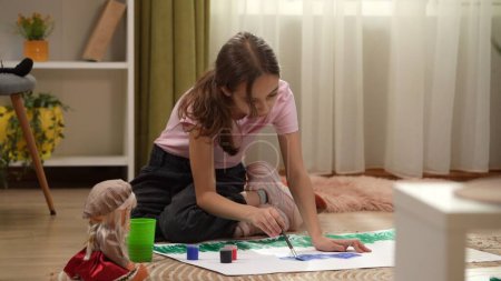 Téléchargez les photos : Dans le cadre, une petite fille est assise par terre, elle tient un pinceau et dessine quelque chose sur du papier. A côté d'elle se trouve une poupée, de la peinture. Elle imagine, fantasme, imagine. Plan moyen. - en image libre de droit