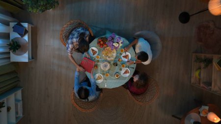 Photo for Family bonding and relationships concept. Scene in the apartment room. Top view of party table with food and drinks. Couple with friends celebrating, wife gives husband birthday present in a box. - Royalty Free Image
