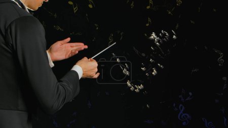 Photo for In a close up shot against a black background, a young man in a tuxedo stands in his hands with a wand. Depicts a conductor who controls the notes, hey surround him and float. He creates art, music - Royalty Free Image