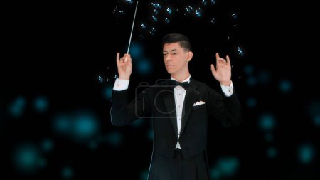 Photo for In a close up shot against a black background, a young man in a tuxedo stands in his hands with a wand. Depicts a conductor who controls the notes, hey surround him and float. He creates art, music. - Royalty Free Image
