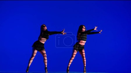 Foto de Dos mujeres bailando tacones bailan en un estudio. Fondo de neón azul, sombra a rayas. Traje sexy negro, tacones altos. Coreografía sensual moderna. Longitud total. Clip promocional o anuncio. - Imagen libre de derechos
