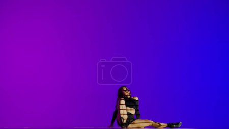 Attractive woman dancing heels dance in a studio. Blue to purple neon gradient background, striped falling shadow. Black sexy costume, high heels. Full length. Promotional clip or advertisement.
