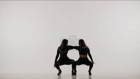 Foto de Dos mujeres bailando tacones bailan en un estudio. Fondo aislado blanco liso. Traje sexy negro, tacones altos. Coreografía sensual moderna. Longitud total. Clip promocional o anuncio. - Imagen libre de derechos