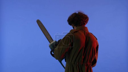 Photo for Man in white mask with chainsaw in hands on blue background with red neon light. Image of maniac Jason Voorhees from Friday 13. View from the back. Halloween celebration concept. - Royalty Free Image