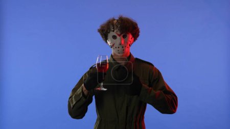 Foto de Hombre con máscara blanca sosteniendo una copa con vino tinto sobre fondo azul con luz de neón rojo. Imagen del maníaco Jason Voorhees del viernes 13. concepto de celebración de Halloween. - Imagen libre de derechos