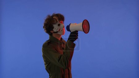 Téléchargez les photos : Homme en masque blanc avec haut-parleur dans les mains sur fond bleu avec néon rouge. Image du maniaque Jason Voorhees du vendredi 13. Concept de célébration Halloween. - en image libre de droit
