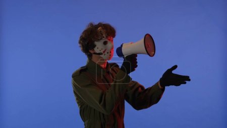 Photo for Man in white mask with loudspeaker in hands on blue background with red neon light. Image of maniac Jason Voorhees from Friday 13. Halloween celebration concept. - Royalty Free Image