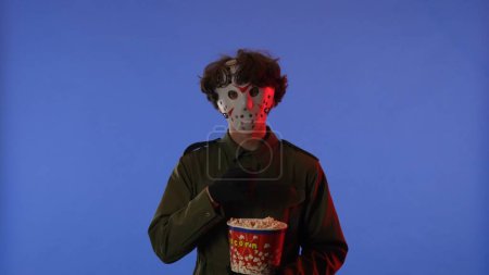 Photo for Man in white mask eating popcorn on blue background with red neon light. Image of maniac Jason Voorhees from Friday 13. Halloween celebration concept. - Royalty Free Image