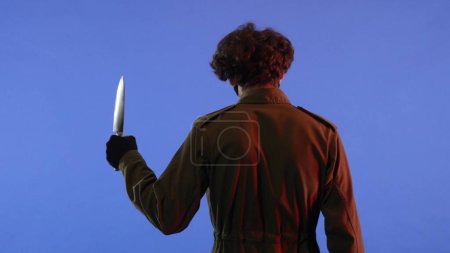 Photo for Man in white mask with sharp big knife in hands on blue background with red neon light. Image of maniac Jason Voorhees from Friday 13. Back view. Halloween celebration concept. - Royalty Free Image