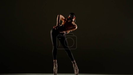 Modern dance style creative advertisement concept. Portrait of female dancer. Appealing woman dancer in high heels standing in extravagant pose at the camera in studio. Isolated on black background.