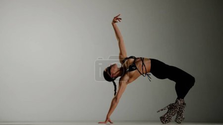 Téléchargez les photos : Style de danse moderne concept publicitaire créatif. Portrait de danseuse. Femme danseuse en talons hauts se tient en pose de pont sur le sol posant dans le studio. Isolé sur fond de dégradé blanc. - en image libre de droit