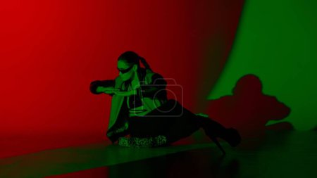 Foto de Concepto de publicidad creativa estilo danza moderna. Retrato de bailarina. Bailarina en tacones altos sentada en el suelo en el estudio con luz de neón roja y verde. Aislado sobre fondo colorido. - Imagen libre de derechos