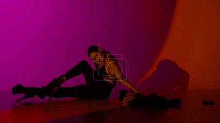 Foto de Concepto de publicidad creativa estilo danza moderna. Retrato de bailarina. Bailarina en tacones altos posando en el suelo en el estudio con luz de neón rosa rojiza. Aislado sobre fondo colorido. - Imagen libre de derechos