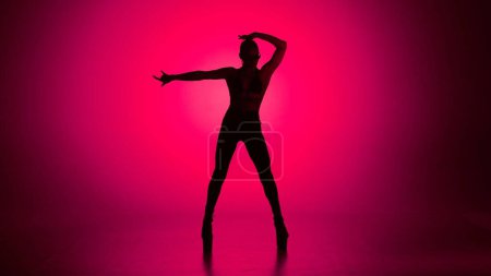 Téléchargez les photos : Style de danse moderne concept publicitaire créatif. Portrait de danseuse. Femme danseuse silhouette en talons hauts debout dans le studio en projecteur rose électrique. Isolé sur fond coloré. - en image libre de droit
