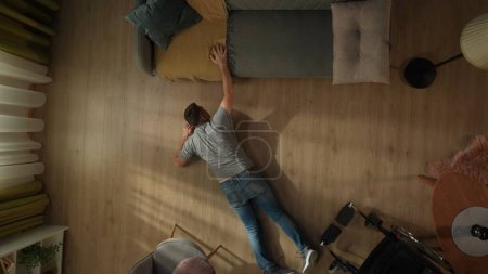 Photo for A disabled man lies on the living room floor after falling from his wheelchair. Top view of a helpless man trying to reach the couch - Royalty Free Image