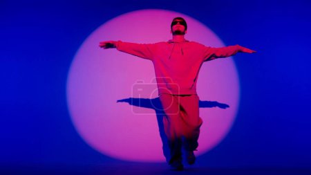 Foto de Un joven se levanta sobre un fondo azul con un foco que le ilumina con un rayo rojo. Lo destaca en un círculo. Demuestra movimiento de baile, posando. Vestido con ropa de calle y gafas. - Imagen libre de derechos