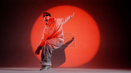 Photo for In the frame on a red background stands a young man on it shines a spotlight beam. Highlights him in a circle. Demonstrates a dance movement, jumping. Dressed in street style clothing and glasses. - Royalty Free Image