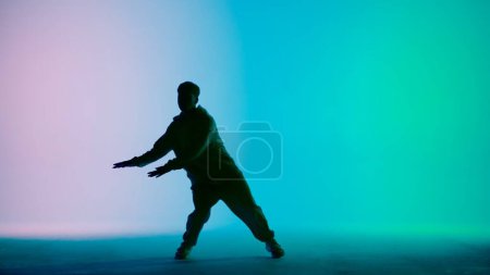 Photo for In the frame on a tricolor background, gradient in silhouette stands a young man. Demonstrates dance moves, stretching his arms forward. He is plastic, rhythmic. He is dressed in street style clothes. - Royalty Free Image