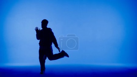 In the frame on a blue and white background, a young man stands in silhouette. Demonstrates dance movements, raising one leg. He is plastic, rhythmic. He is dressed in street style clothes. Medium.