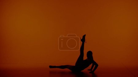 Photo for In the frame on a red background. Sits a woman in silhouette, she is wearing revealing clothing and long heels. Demonstrates a dance movement, pose, stretching. Raising one leg up. She is sexy. - Royalty Free Image
