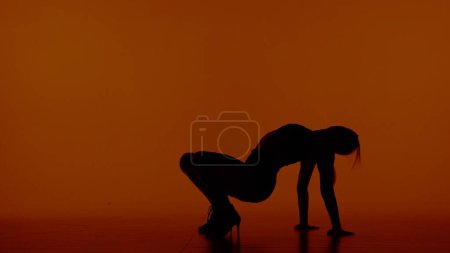 Téléchargez les photos : Dans le cadre sur un fond rouge, orange. Une femme en silhouette porte des vêtements ouverts et des talons hauts. Démontre un mouvement de danse et pose. Elle s'assit les mains sur le sol. Shes sexy. - en image libre de droit