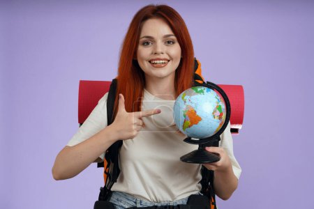 Photo for Tourism and active leisure time advertisement concept. Portrait of female traveler. Woman tourist in casual with backpack holding globe and pointing at the map, smiling. Isolated on purple background. - Royalty Free Image