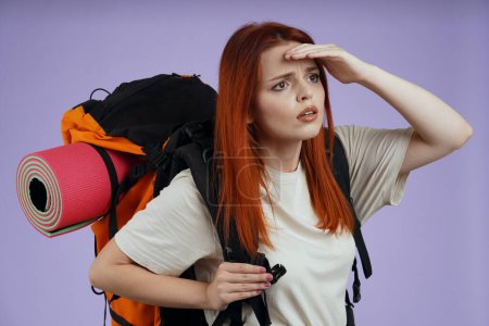 Photo for Tourism and active leisure time advertisement concept. Portrait of female traveler. Woman tourist in casual with backpack holding hand at forehead looking at the side. Isolated on purple background. - Royalty Free Image