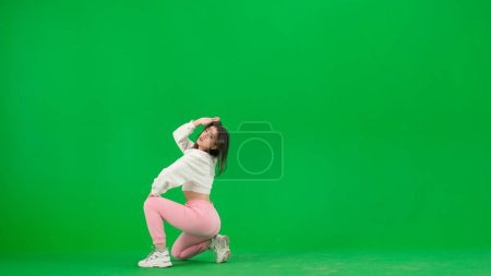 Téléchargez les photos : Chorégraphie de danse moderne concept publicitaire créatif. Jolie femme en haut blanc et pantalon rose assis dans la pose de jazz funk danse sur fond chroma écran vert clé dans un studio. - en image libre de droit