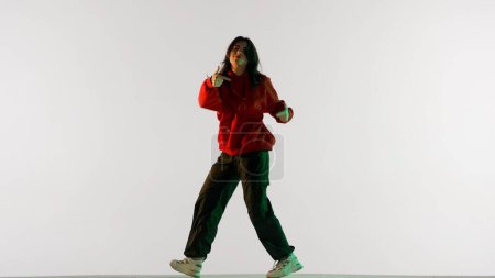Foto de Coreografía de danza moderna concepto de publicidad creativa. Mujer atractiva en sudadera con capucha roja y pantalones negros bailando jazz funk, aislado sobre fondo blanco iluminado por luz de neón verde. - Imagen libre de derechos