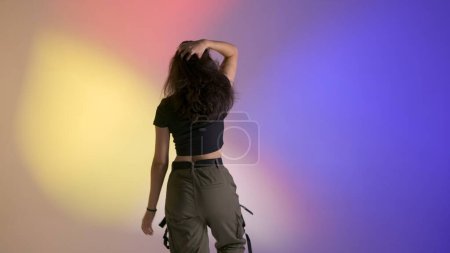 Photo for Modern dance choreography creative advertisement concept. Attractive woman in top and khaki pants posing with her back at camera close up shot, dancing jazz funk in studio, isolated on neon background - Royalty Free Image