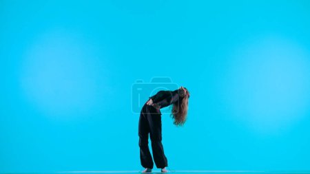 Photo for Modern creative choreography and dance concept. Portrait of female dancer. Attractive girl silhouette in black clothing showing contemporary dance elements on blue background in studio. - Royalty Free Image