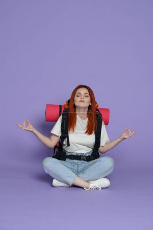 Photo for Tourism and active free time advertisement concept. Portrait of girl traveler. Woman tourist in casual with backpack sitting on the floor hands in meditative position. Isolated on purple background. - Royalty Free Image